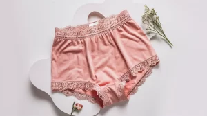 Calcinha boxer feminina rosa pastel com renda delicada em fundo branco, ao lado de uma pequena decoração floral.