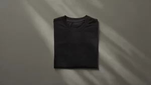 Camiseta masculina preta de algodão dobrada em um fundo cinza neutro, com reflexos de luz destacando detalhes do tecido.